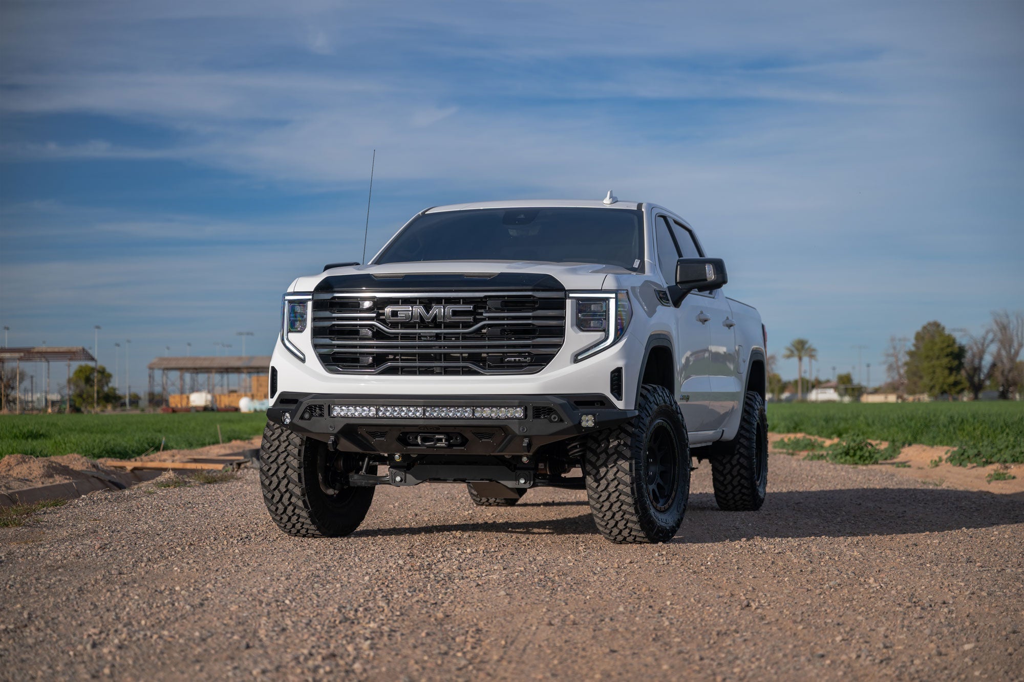 White GMC Sierra 1500 with Stealth Fighter Front Bumper and Winch Kit Upgrade on a dirt road with a clear sky
