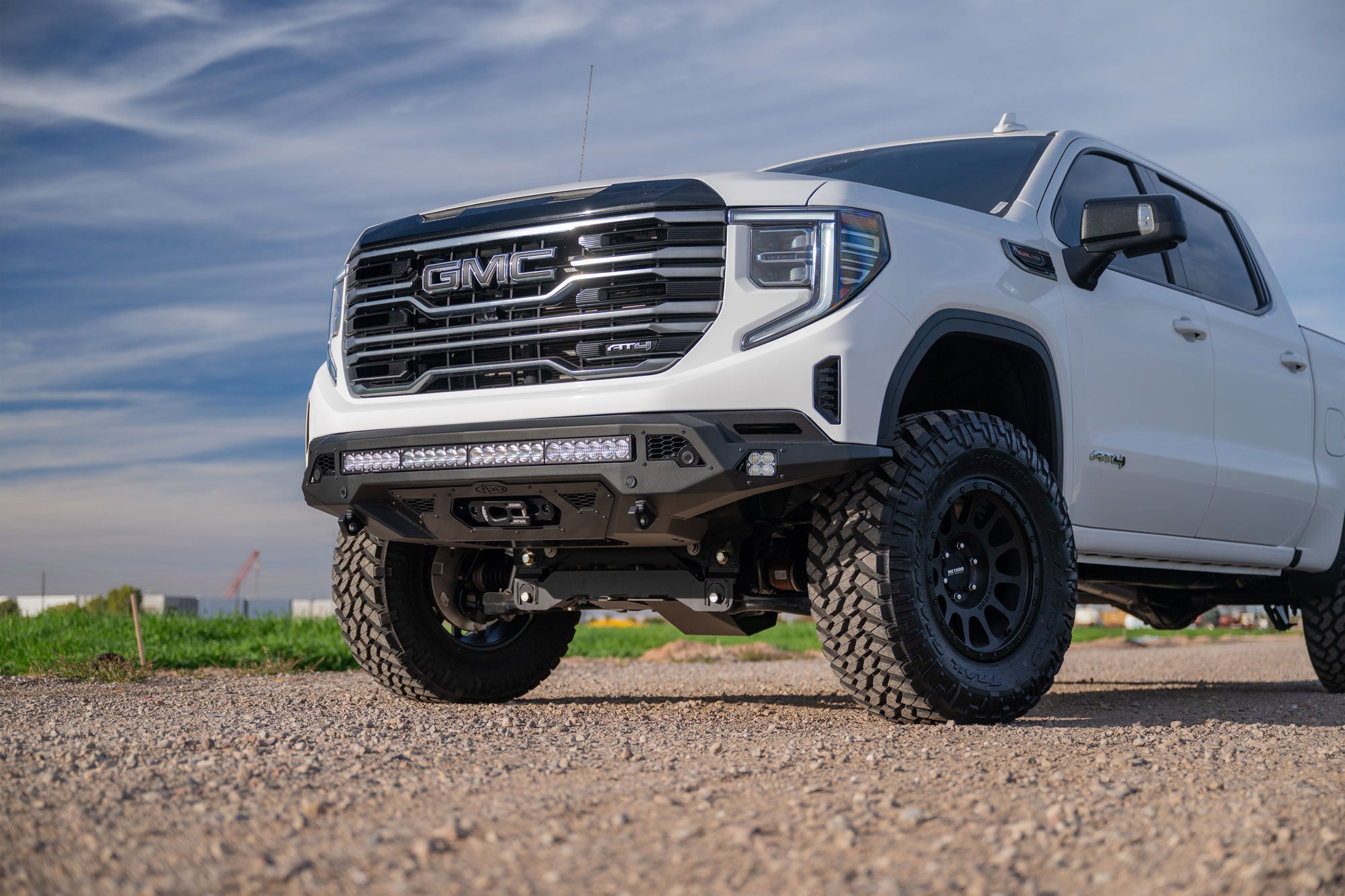 White GMC truck with Stealth Fighter Front Bumper and Winch Kit Upgrade on a gravel road under a blue sky.