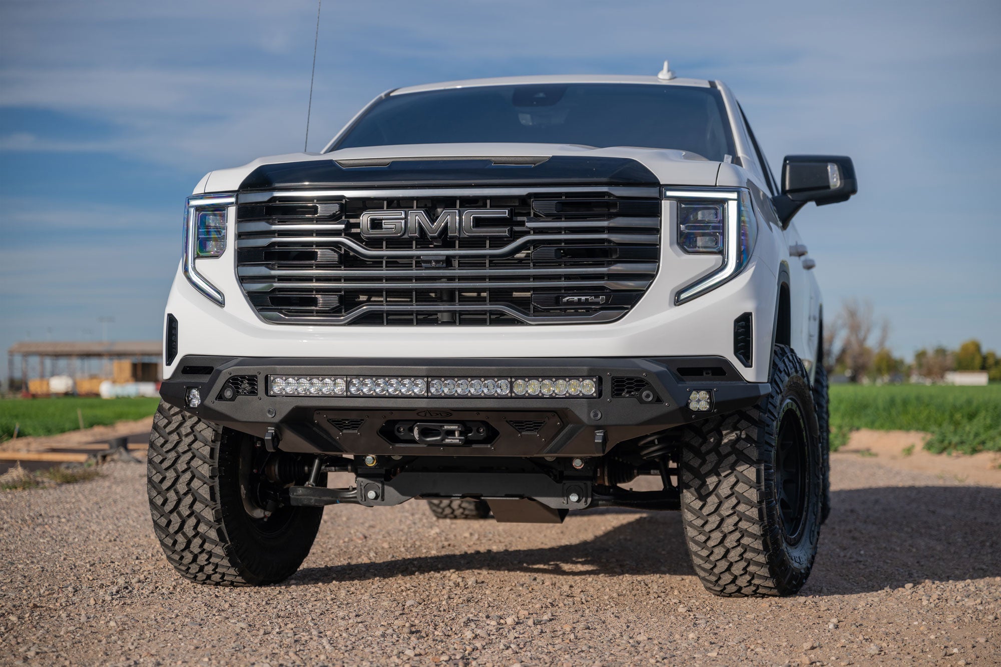 White GMC Sierra 1500 with Stealth Fighter Front Bumper and Winch Kit Upgrade on a gravel road with a clear sky background