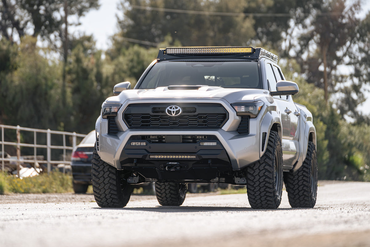A silver Toyota Tacoma pickup truck with a custom black ADD Center Mount front bumper, off-road tires, roof rack, and light bar, photographed on a gravel road with trees in the background.