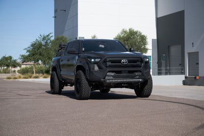 Black Toyota truck parked on a paved surface with industrial buildings in the background. Stealth Fighter Winch Front Bumper for the 4th Gen Toyota Tacoma installed.
